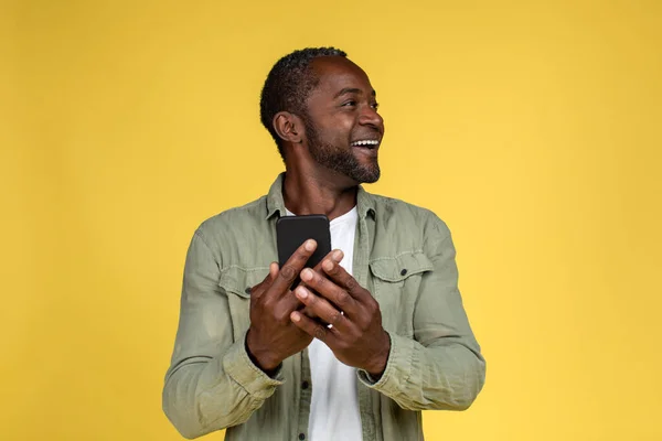 スマートフォンで入力し 黄色の背景 スタジオに隔離された空白のスペースを見ている大人の黒人男性を笑顔 感情や表情 仕事やビジネスのためのガジェットをリモートで アプリとオファー — ストック写真
