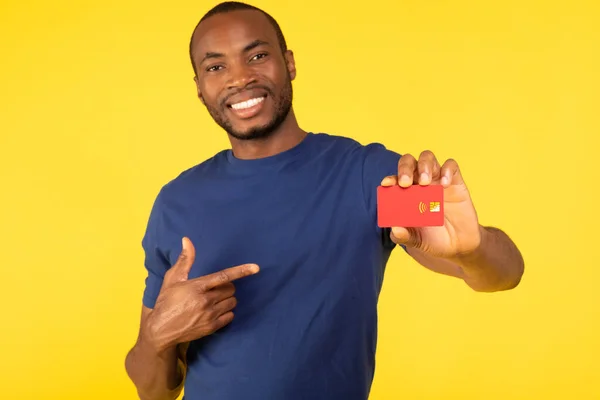 カメラ広告に赤いクレジットカードを示すあなたの黒い男偉大な銀行のオファー黄色のスタジオの背景に指を指しています 金融と銀行サービスの概念 選択的フォーカス — ストック写真