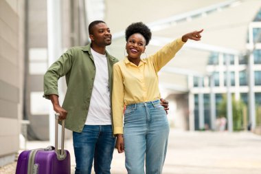 Harika bir seyahat teklifi. Çantalı mutlu siyah turist çifti, havalimanında birlikte seyahat ederken parmağıyla işaret ediyor. Turizm ve Ulaştırma Konsepti