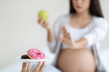 Yeşil elma tutan, çöreklerle dolu tabağı reddeden ve sağlıksız yiyeceklere hayır diyen bir bayanı bekleyen tanınmamış hamile bir kadın. Sağlıklı beslenme alışkanlıkları hamilelik ilaçlama