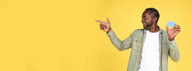 Açık ağızlı, kendine güvenen siyah bir erkek. Kredi kartı, sarı arka planda, stüdyoda izole edilmiş boş bir yeri işaret ediyor. Nakit para, harika indirim, online alışveriş, reklam ve teklif