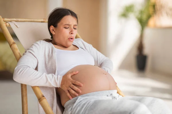 Pregnancy Pain. Young Pregnant Woman Touching Belly While Sitting In Chair At Home, Worried Expectant Female Suffering Painful Contractions Or Braxton Hicks, Making Breathing Exercises To Relief Ache