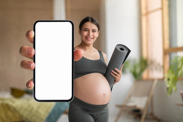 Pregnant Woman With Fitness Yoga Mat In Hand Demonstrating Big Blank Smartphone With White Screen At Camera, Happy Young Expectant Lady Recommending New Pregnancy App Or Website, Mockup