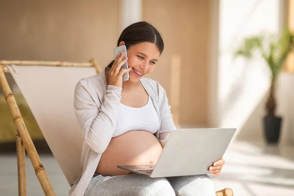 怀孕期间的自由 年轻的怀孕妇女与笔记本电脑一起工作 坐在椅子上用手机交谈 享受远程工作的快乐期待 复制空间 — 图库照片