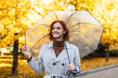 Bin yıllık kızıl saçlı, şemsiyeli neşeli beyaz kadın sonbaharda sarı yapraklarla parkta yağmurun tadını çıkarıyor. Dışarıda tek başına yürümek, iyi bir ruh hali ve şehirde yaşam tarzı.
