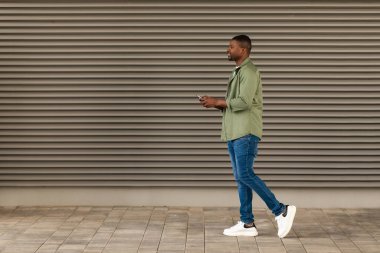 Siyah adam cep telefonu mesajlaşması kullanıyor. Çevrimiçi iletişim kuruyor. Dışarıya doğru yan yan bakıyor. Erkek Kullanımı Harika Uygulama ve Dışarıda Mobil Oyunlar Oynuyor. Teknoloji ve Gadget. Tam Uzunluk