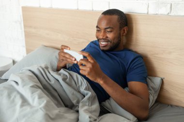 Neşeli Afrikalı Amerikalı adam cep telefonu kullanıyor. Mobil oyun oynuyor. Yatakta uzanıyor, modern yatak odasında eğleniyor. Smartphone için Harika Uygulama