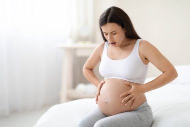 Braxton Hicks contractions. Young pregnant woman in pain suffering from abdominal ache at home, sitting on bed, touching her big tummy, having cramp, panorama with free space clipart
