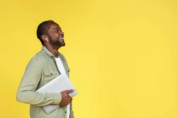 Laughing adult african american guy in wireless headphones with laptop looking up at empty space, isolated on yellow background, studio. Technology for study, work and business, good ad and offer