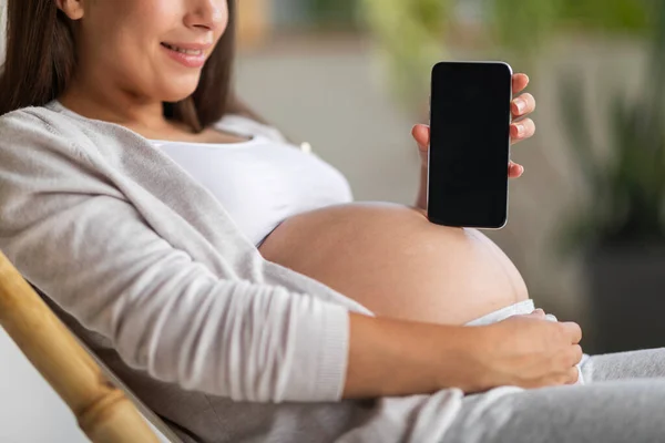 Young Pregnant Woman Holding Blank Smartphone While Relaxing In Chair At Home, Unrecognizable Expectant Lady Embracing Her Belly And Showing Empty Cellphone With Black Screen, Cropped, Mockup