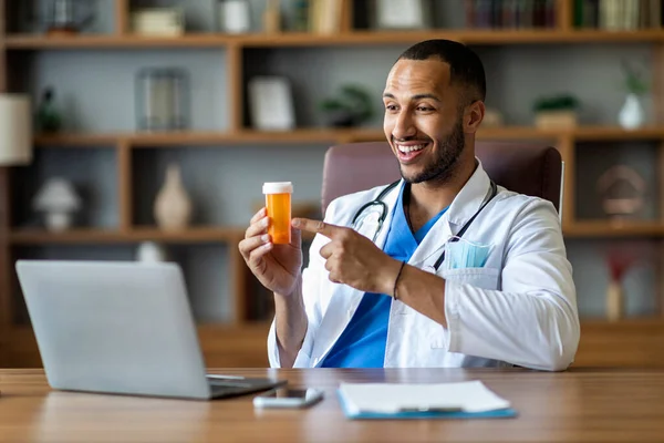 Friendly young black man in workwear doctor working on computer at clinic, looking at laptop screen, showing medication and smiling, having video conference with patient, copy space. Remote medicine