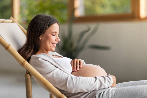 Beautiful smiling pregnant woman hugging her belly while relaxing in chair at home, young expectant lady embracing tummy and resting in living room, enjoying pregnancy time, side view, copy space