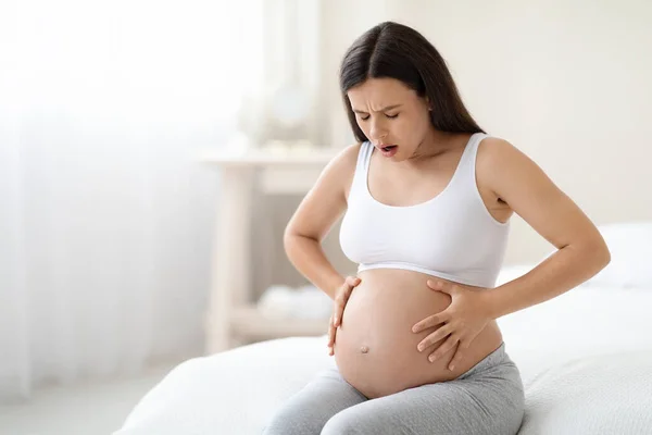 Braxton Hicks contractions. Young pregnant woman in pain suffering from abdominal ache at home, sitting on bed, touching her big tummy, having cramp, panorama with free space