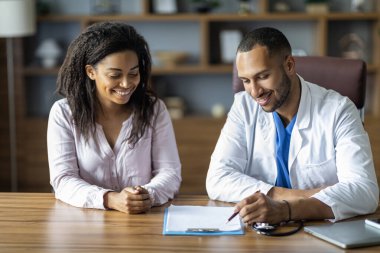 Neşeli Afro-Amerikalı yakışıklı erkek doktor ve güzel kadın hasta modern klinikte masada oturmuş sohbet ediyorlar, doktor pratisyen kadın danışman.