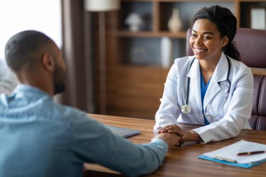 Çekici Afrikalı Amerikalı genç kadın doktor, hasta adamın elini tutuyor ve gülümsüyor. Genel pratisyen, modern klinikteki danışmanlık sırasında hastayı rahatlatıyor.