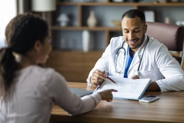 Doktor konseptiyle randevu. Beyaz giysili yakışıklı Afro-Amerikan adam hasta zenci kadın tedavi planı gösteriyor, terapist ve kadın konuşuyor, klinik içi.