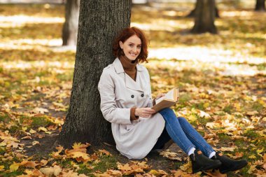 Bin yıllık mutlu, kızıl saçlı, yağmurluklu kadın kitap okuyor, parkta sarı yapraklarla oturuyor, boş yer var. Biraz ara ver, edebiyat hobisi, dinlenme ve rahatlama şehirde tek başına.