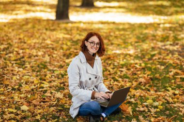 Bin yıllık kızıl saçlı neşeli bir kadın ve dizüstü bilgisayarında yazan gözlüklü, sarı yapraklarla parkta oturuyor. Dinlen ve dışarıda dinlen, sosyal ağlarda, işte ve işte sohbet et.