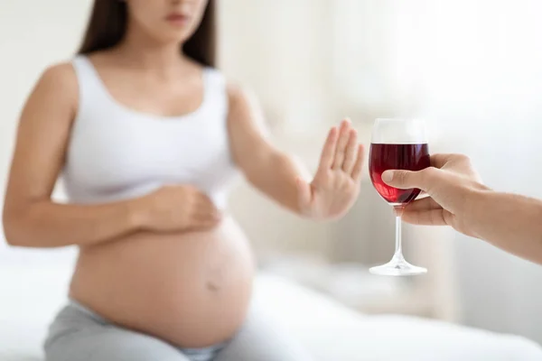 No alcohol during pregnancy. Unrecognizable young pregnant woman refuses to drink wine, making stop gesture to glass, sitting on bed at home, touching her belly, free space, cropped shot