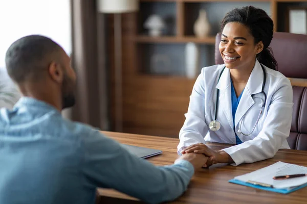 黒人男性患者の手と笑顔を保持する魅力的なアフリカ系アメリカ人の若い女性医師 現代の診療所での協議中に患者を慰め一般開業医 コピースペース — ストック写真