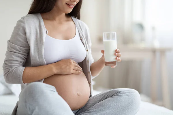 Cropped of expecting lady with big tummy sitting on bed at home, holding glass, unrecognizable pregnant woman drinking milk, white bedroom interior, copy space. Calcium for baby growth concept