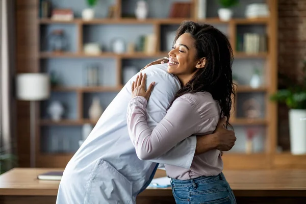 Doctor unrecognizable man in white workwear hugging cheerful young black lady patient, celebrating good news, clinic interior, copy space. Doctor and patient relationships concept