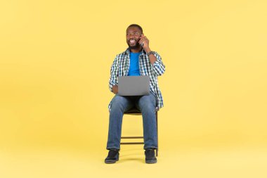 Cep telefonuyla konuşan, internetten konuşan, sarı arka plan üzerinden stüdyoda oturan mutlu Afrikalı Amerikalı adam. İnternet Teknolojisi ve İletişim