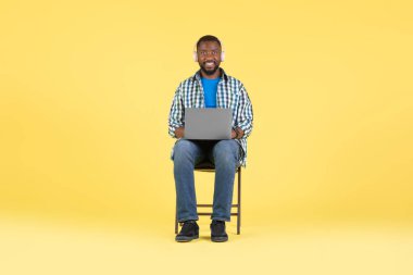 Laptop kullanan, kablosuz kulaklık takan, müzik dinleyen ya da internette öğrenen sarı arka planda sandalyede oturan, kameraya gülümseyen Afrikalı Amerikalı bir erkek. Teknoloji ve İnternet