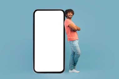 Havalı yakışıklı Hintli adam beyaz ekranlı büyük cep telefonuna yaslanıyor, kolları çapraz, mavi stüdyo arka planı, mobil uygulama veya reklam için model, kopyalama alanı