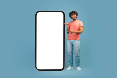 Neşeli, kıvırcık, günlük kıyafetli, beyaz ekranlı akıllı telefonu işaret eden, gülümseyen, iyi bir mobil teklif öneren, mavi stüdyo geçmişi olan,