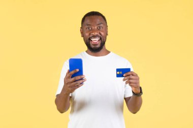 Mobil Alışveriş Uygulamasını Akıllı Telefon ve Kredi Kartıyla Sarı Stüdyo Arkaplanında Ödeme Yapan Kameraya Gülümseyen Mutlu Siyah Adam. Ticaret, Banka Uygulaması Konsepti