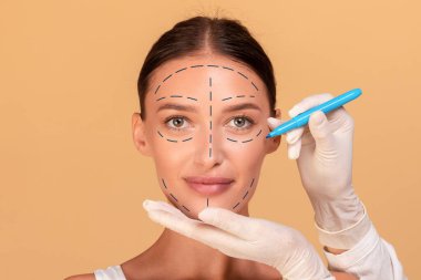 Estetik ameliyat kavramı. Genç bayan kozmetoloji kliniğinde görünüşünü değiştiriyor. Plastik cerrah, güzel kadın yüzüne izler bırakıyor. Bej rengi stüdyo arka planı.