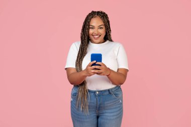 Mutlu Siyahi Kadın, Stüdyoda Pembe Arkaplanda Duran Kameraya Gülümseyen Telefon Mesajları Yazıyor. Cep telefonu reklamları yapan şişman kadın pozu. Mobil uygulaması. Teknoloji ve Malzemeler