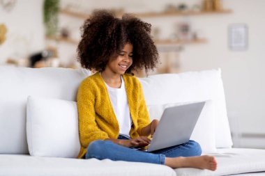 Mutlu Afro-Amerikan çocuk küçük kız evdeki kanepede oturuyor, modern laptop kullanıyor, bilgisayar klavyesinde daktilo kullanıyor, bilgisayarlarda eğitim oyunları oynuyor, yer kopyalıyor.