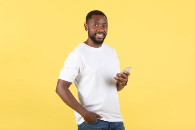 Sarı Stüdyo Arkaplanının Üzerinde Duran Sosyal Medya Habercisi aracılığıyla Kameraya Gülümseyen Cep Telefonu Mesaj Posteri 'ni kullanan Afro-Amerikalı Mutlu Noeller. Kaynaklar ve Teknoloji