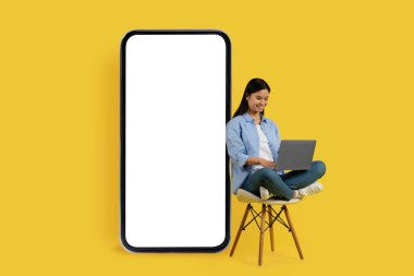 Gülümseyen genç Çinli kız öğrenci, sarı arka planda izole edilmiş, boş ekranlı, büyük bir akıllı telefonun yanında dizüstü bilgisayarla koltukta oturuyor. Uzaktan eğitim uygulaması, dijital göçebe, reklam ve teklif