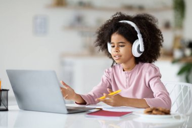 Çalı saçlı zeki siyah kız öğretmeniyle video görüşmesi yapıyor, evde dizüstü bilgisayarın önünde oturuyor, kablosuz kulaklık kullanıyor, bilgisayar ekranına bakıyor, el kol hareketi yapıyor ve gülümsüyor.