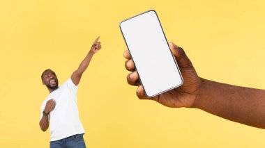 Sarı Stüdyo Arkaplanı 'nda Wow uygulaması pozu öneren boş ekranlı, dev cep telefonuyla işaret eden heyecanlı siyah adam. Büyük Mobil Teklif Konsepti. Panorama, Mockup
