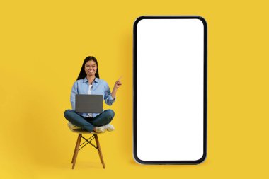 Genç Koreli kız öğrencinin dizüstü bilgisayarla sandalyede oturup boş ekranlı akıllı telefonu göstermesi, sarı arka planda ve stüdyoda izole edilmiş yeni bir uygulama önermesi güzel. Uzak ve yeni normal çalış