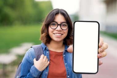 Eğitim Reklamı. Gülümseyen Arap Kız Öğrenci Açık havada dururken Büyük Boş Akıllı Telefonu Gösteriyor, Gözlüklü Genç Orta Doğulu Kadın Kamerada Beyaz Ekran ile Cep Telefonu Gösteriyor, Mockup