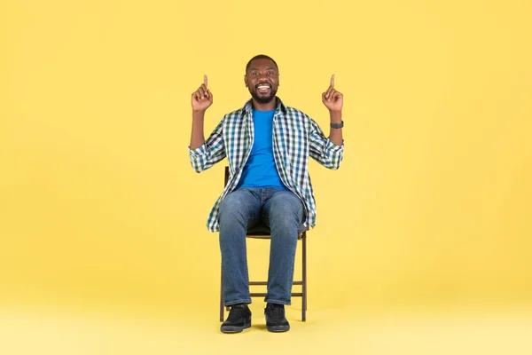 图上的非洲裔美国人双手高举指尖 坐在椅子上 在黄色的工作室背景上刊登广告 微笑着拍照 向上看概念 — 图库照片