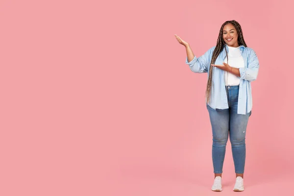 Oversized Black Female Pointing At Free Space For Text Smiling To Camera Standing Over Pink Studio Background, Advertising Great Offer. Look There. Full Length Shot