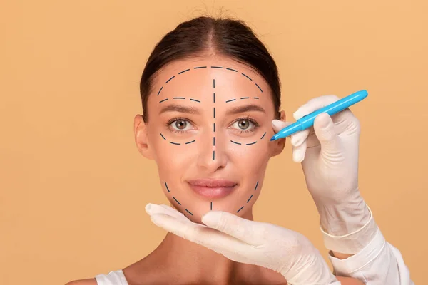 Concept of plastic surgery. Young lady changing her appearance at cosmetology clinic, plastic surgeon making marks on beautiful woman face, beige studio background