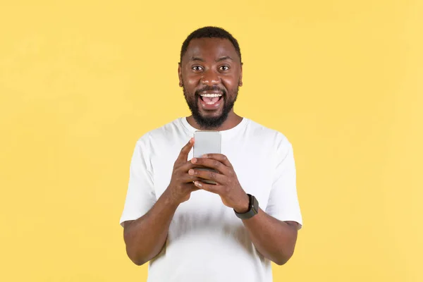 兴奋的黑人男性使用手机微笑拍摄在兴奋的拍摄黄色工作室背景 家伙发短信和使用伟大的应用程序在手机上 哇移动提供概念 — 图库照片