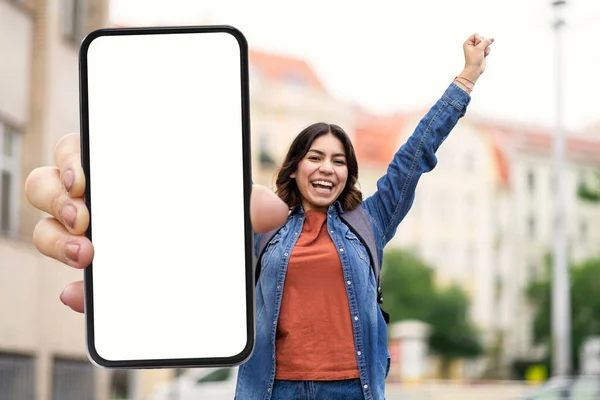 Mobile Ad. Happy Arab Female Student Showing Big Blank Smartphone With White Screen And Celebrating Success While Standing Outdoors, Cheerful Young Woman Recommending New App Or Website, Mockup