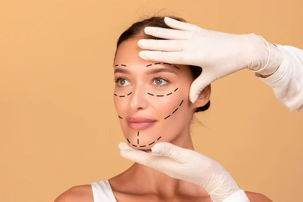 Frontal portrait of young woman getting spa treatment from beautician in protective gloves. Plastic surgeon making marks on female face over beige studio
