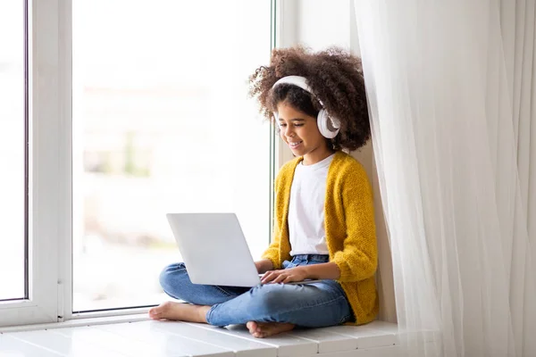 自宅でラップトップを使用してワイヤレスヘッドフォンでかわいい黒の女の子は ウィンドウに座って 画面を見て コンピュータのキーボードを入力し コンピュータ上でビデオゲームをプレイし スペースをコピー — ストック写真