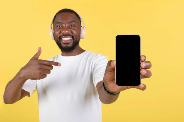 無線イヤフォンを装着した携帯電話のブランク画面を表示する幸せなアフリカ系アメリカ人男性スタジオで黄色の背景にある携帯電話で指を指す音楽アプリケーションを宣伝します 選択的フォーカス — ストック写真