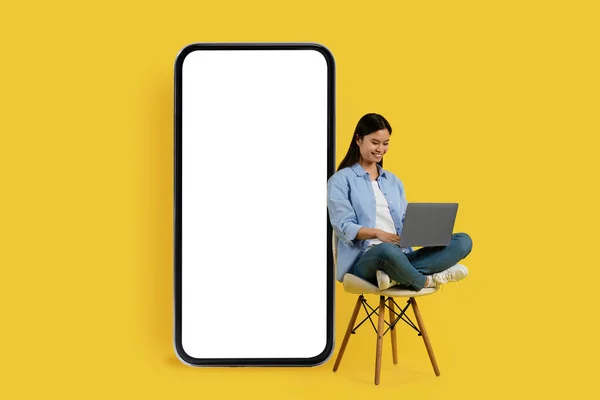 Smiling busy pretty young chinese lady student sits on chair with laptop near huge smartphone with blank screen, isolated on yellow background. App for study remote, digital nomad, ad and offer