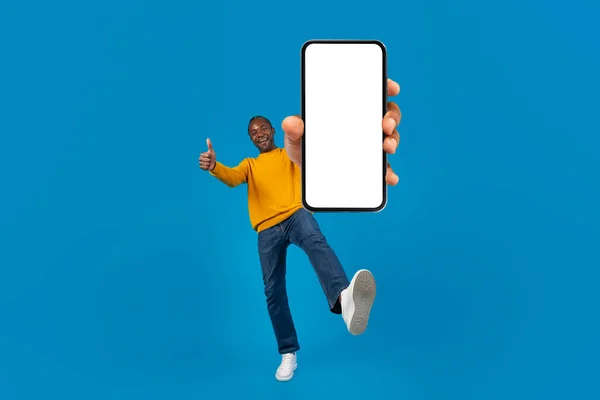 Cheerful mature black man in casual showing brand new cell phone with white blank screen and gesturing thumb up, lifting leg up, blue studio background, panorama with copy space, mockup, full length